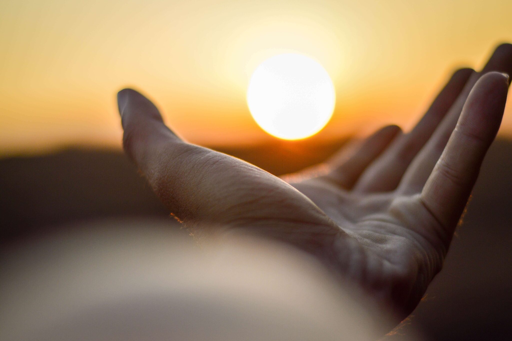 eine Hand im Vordergrund hält optisch täuschend die Sonne im Sonnenuntergang in der Hand, die Hand im Vordergrund ist scharf, der Rest des Bildes ist unscharf