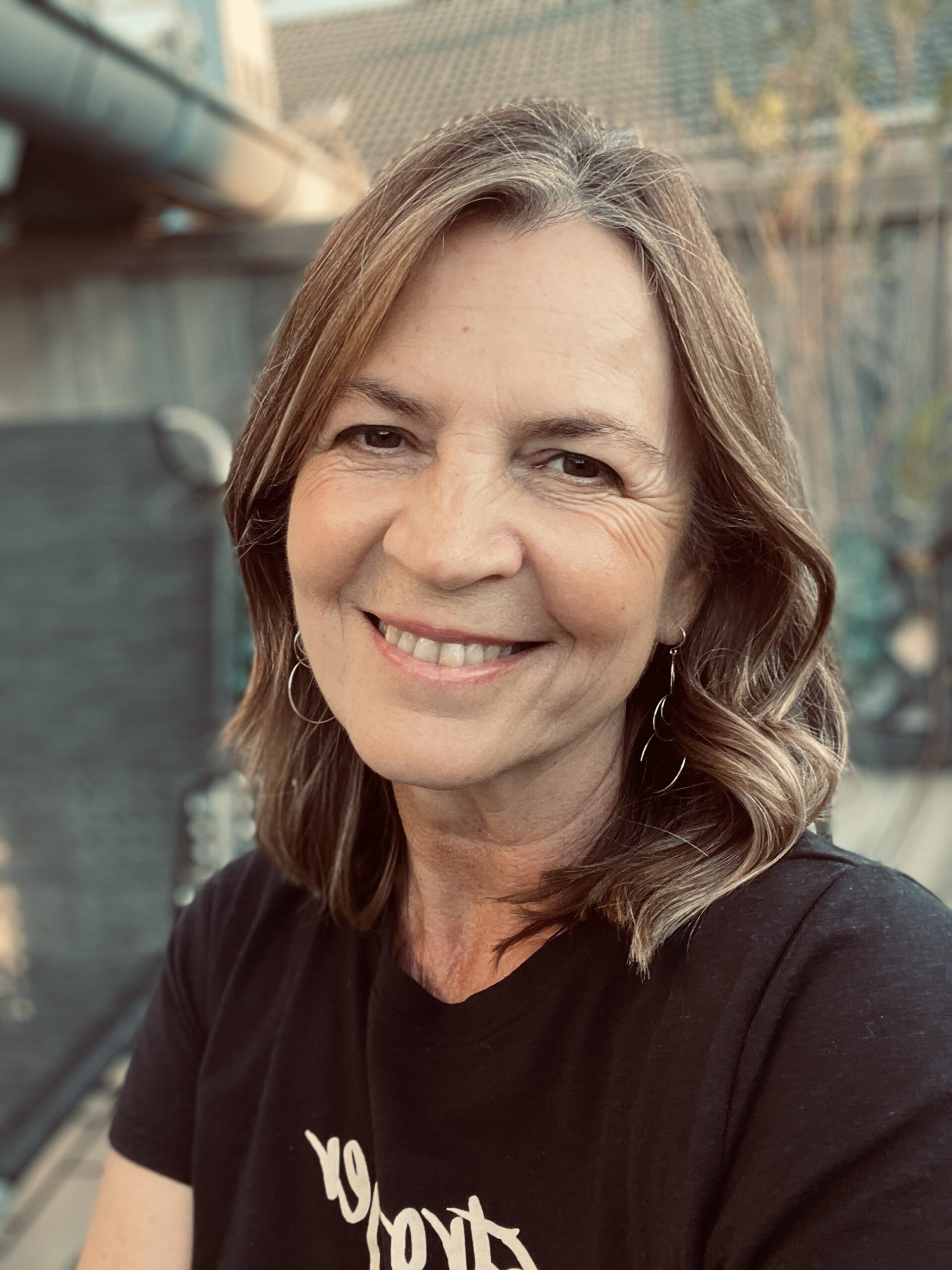 Karin sitzt im Freien am Balkon , trägt ein schwares Kurzarmshirt un dlächelt in die Kamera.